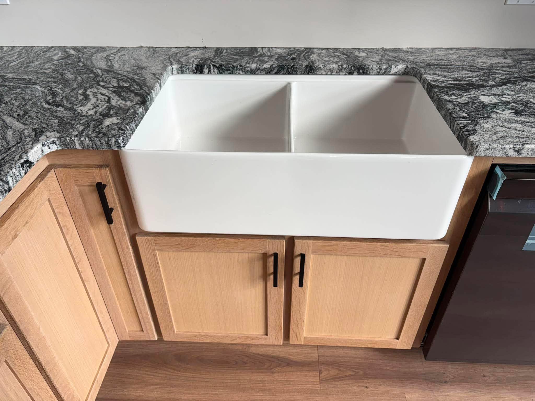 White porcelain double-bowl farmhouse sink set into dark granite countertop, natural oak sink base cabinet below.