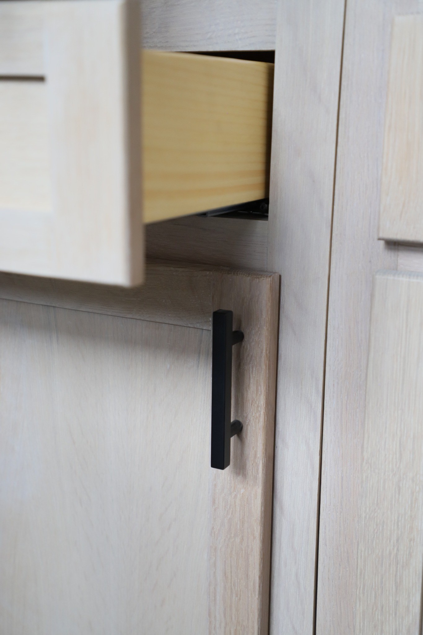 Matte-black cabinet pull mounted on a natural wood shaker-style door.